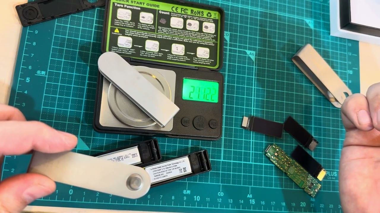 ภาพจากคลิป Weight and Ledgers ของ Mark Karpelès ฝาโลหะของ Ledger Nano X วางบนตาชั่งดิจิทัลแบบละเอียด หน้าจอแสดง 21.122 ด้านล่างเป็นแบตเตอรี่ 100mAh ที่แยกออกมา